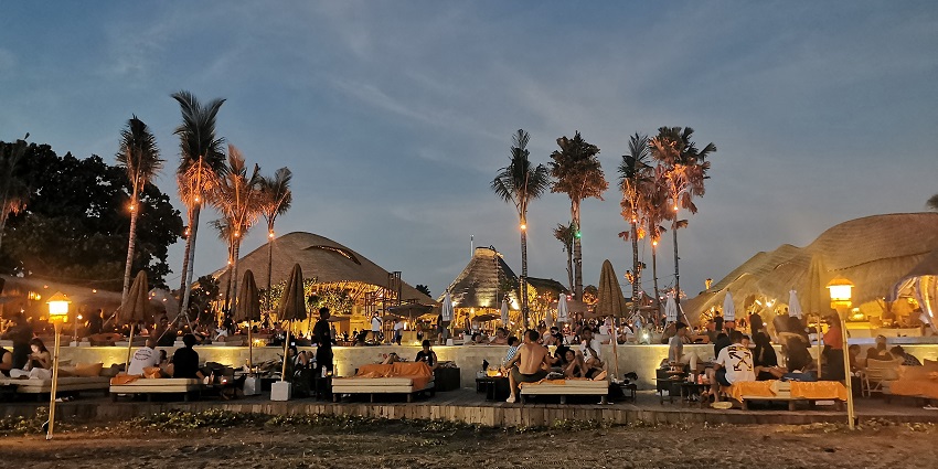Open-air beach club at night with people sitting on lounge chairs and palm trees