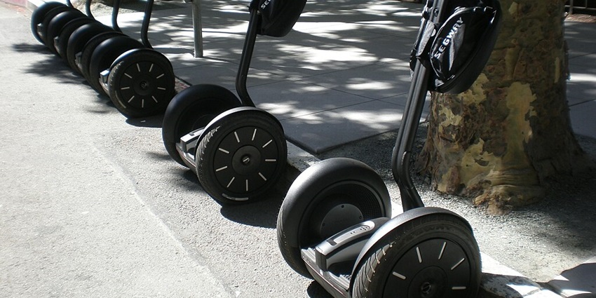 Several segways parked under a tree which is outside a park in front of its gates.