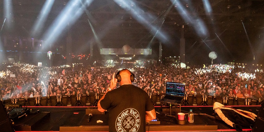 A DJ wearing sunglasses and a hat performing at a nightclub party, surrounded by an energetic crowd.
