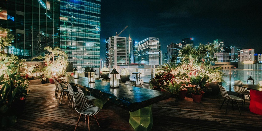 A rooftop dining area at night with a long table, chairs, plants, and a backdrop of city