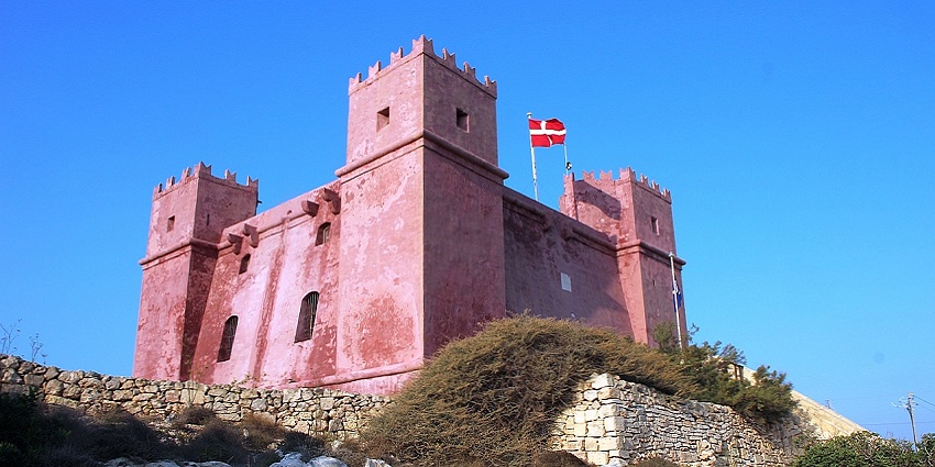A red-coloured tower is standing on a highland surrounded by lush green on a sunny day.