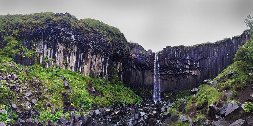 An Image of water flowing from the Svartifoss Waterfall, surrounded by greenery and rocky cliffs.