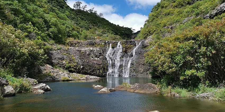 Tamarind waterfall flowing into the calm river, surrounded by greenery against blue sky.