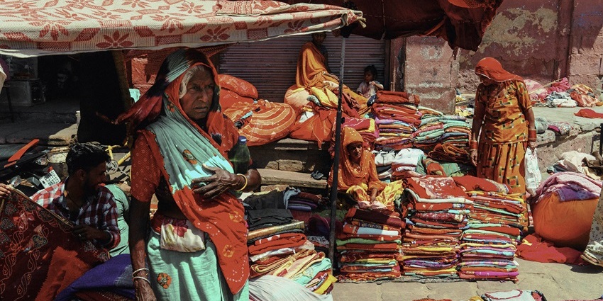 A picture of an old woman in a shopping street in Ayodhya with multiple cloth stalls in background.
