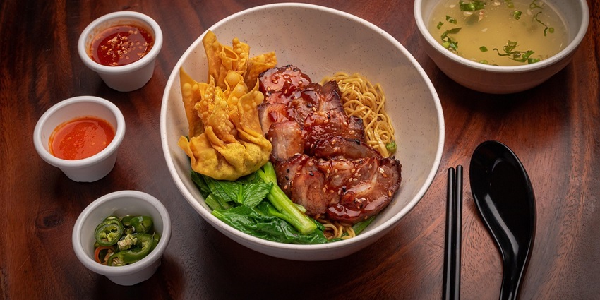 Noodles served with veggies and fried wontons in a bowl alongside soup and chillies.