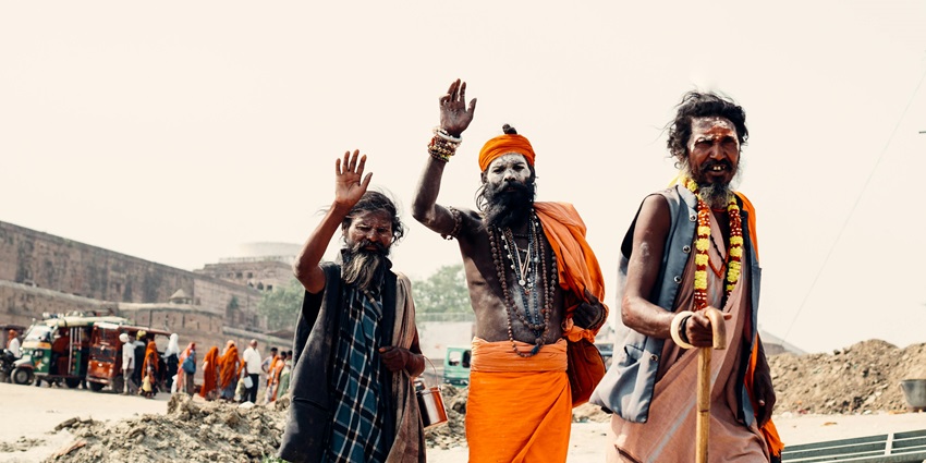 Three Naga Sadhus in saffron-coloured clothes are sitting in front of a wooden door.