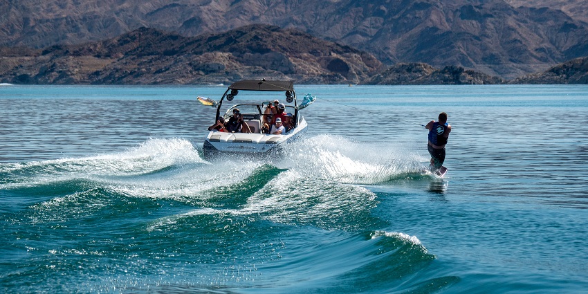 People on a boat in Lake Mead with a person wakeboarding creating waves and hills visible