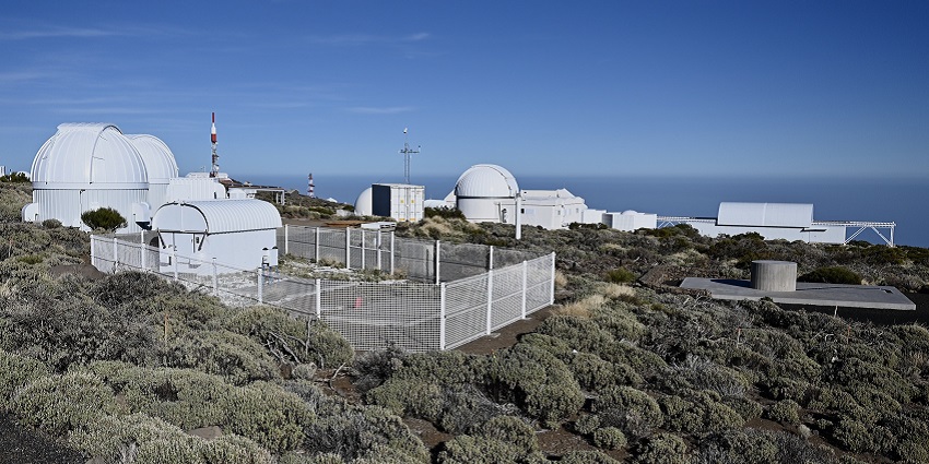 An image of a solar observatory atop a hill, surrounded by trees, under a clear blue sky.
