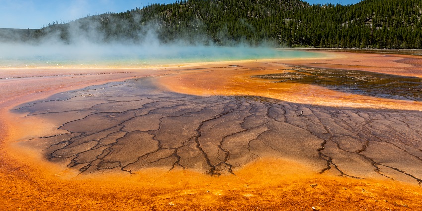A colourful Grand Prismatic Spring with rugged ground and smoke erupting and greenery in the background