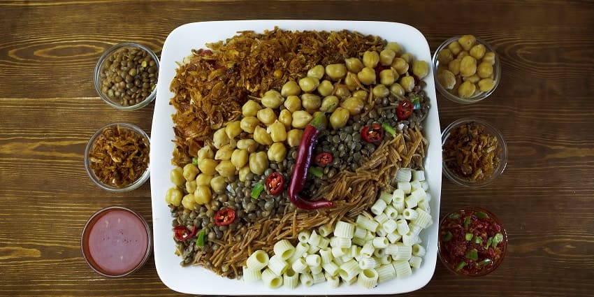 A plate filled with Egyptian koshari is presented with a few food items on the side