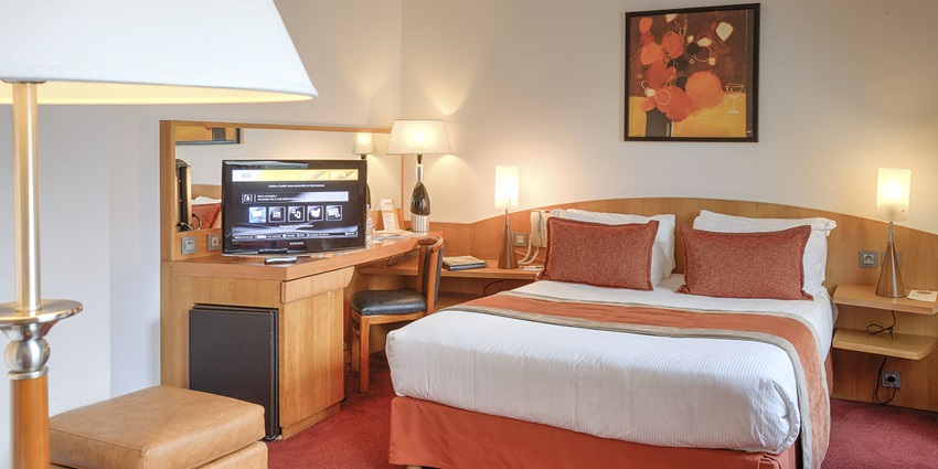 A well-lit hotel room that has a cozy bed, desk, lamp, and all other necessities