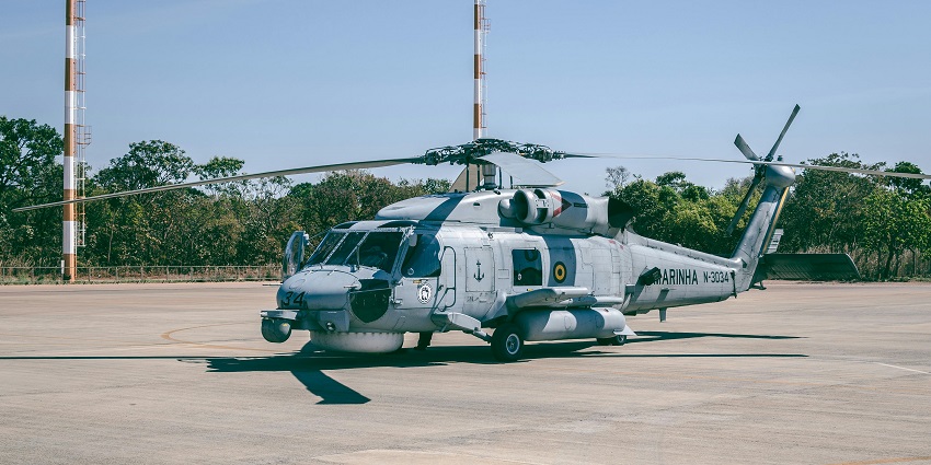 Grey helicopter parked at an airstrip with tall red and white iron poles and greenery.