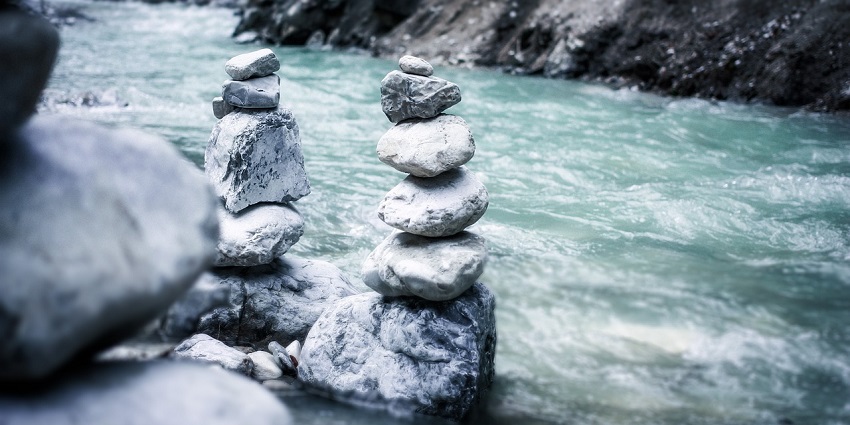 Stacked river rocks balanced on a large stone by a calm river surrounded by nature.