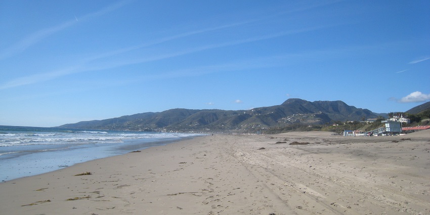 Wide, expansive Zuma Beach, with a small hill on the backdrop, is empty on a sunny day