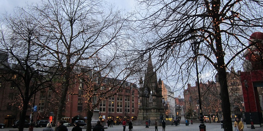 Manchester's Albert Square with its gothic buildings, monuments, and people gathered around.