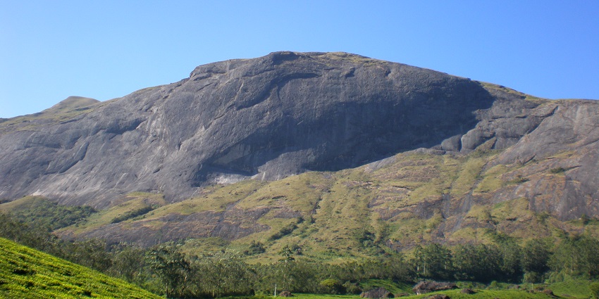 An image of Anamudi Peak, a large rocky mountain with a green grassy foreground under a clear blue sky.