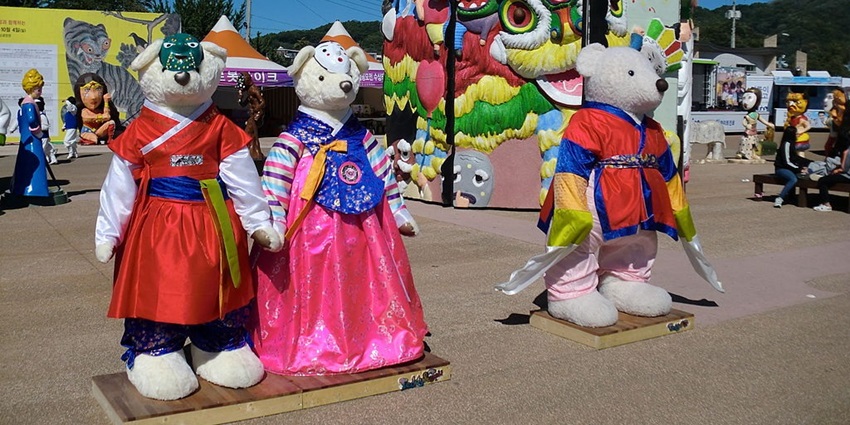 Teddy bears in Korean clothing at a festival, with a colorful, folk-art inspired backdrop