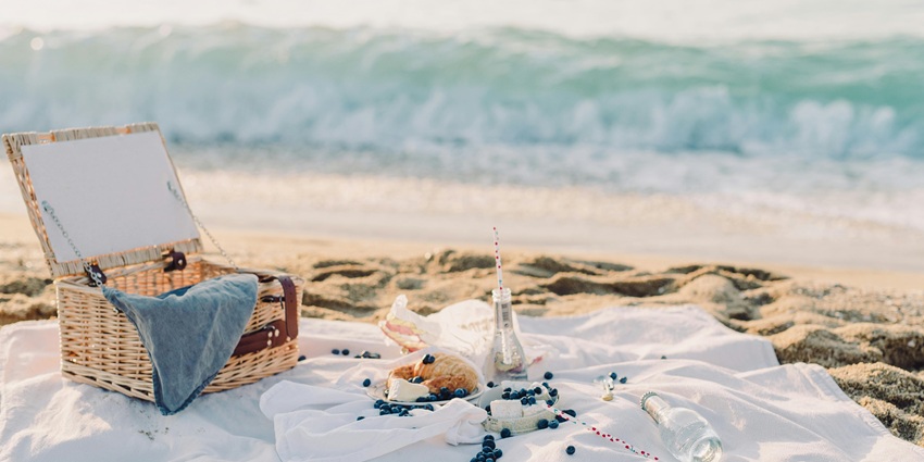 A picnic basket, blanket, and snacks laid out on a sandy beach with waves in background