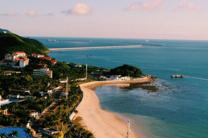 An image of a beach in China, with green hills, and a long pier extending into the ocean.