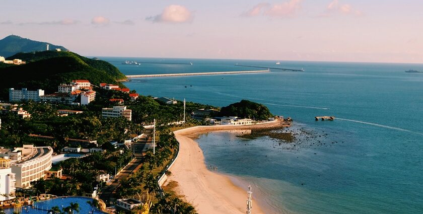 An image of a beach in China, with green hills, and a long pier extending into the ocean.