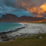 An image of a beautiful beach in Norway with clear blue waters and a backdrop of stunning mountains.