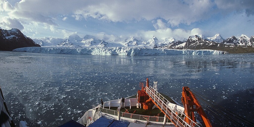 A ship's bow points towards a massive glacier calving into the ocean, with snowy mountains