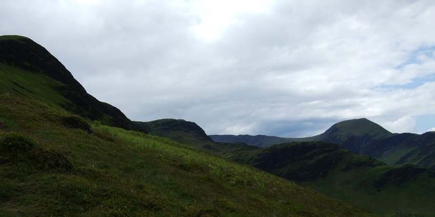 A grassy hillside slopes down into a valley surrounded, green mountains under a cloudy sky