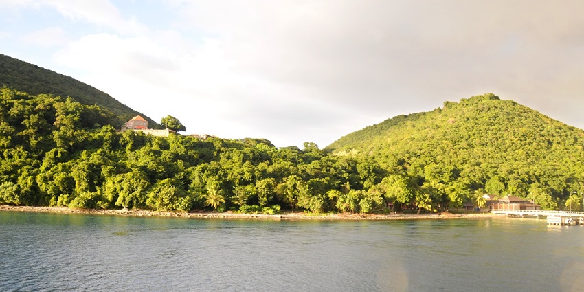 Panoramic view of the Cabrits National Park between two hills filled with greenery and a structure on top.
