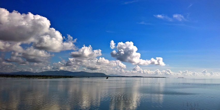 Chilika Lake with a still water body and distant mountains under the blue cloudy sky.