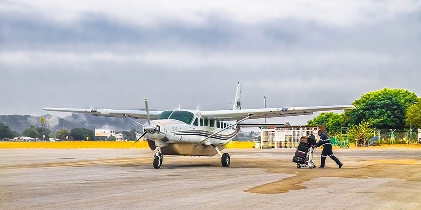 A small plane is standing on the Dodoma airport Tarmac, and a man walks towards it with a trolley full of bags