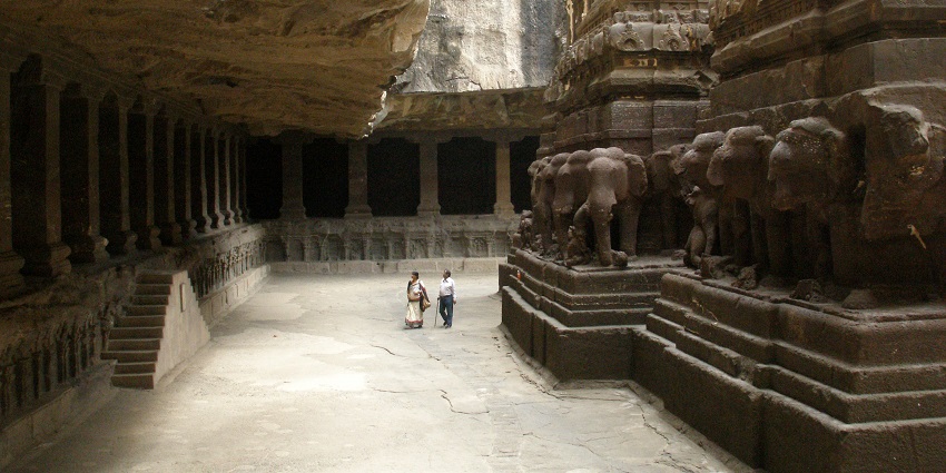  An image of two people walking inside an ancient rock-cut temple with intricate carvings and elephant sculptures
