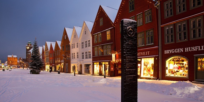 An image of Bryggen in Bergen, Norway, with snow-covered ground and illuminated colorful wooden buildings at dusk.