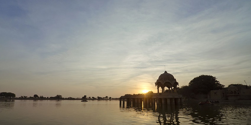An image of Gadsisar Lake in Rajasthan at sunset with historic structure, and calm waters