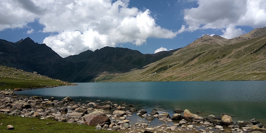Gangabal Lake with a rocky shoreline surrounded by green mountains under a cloudy sky