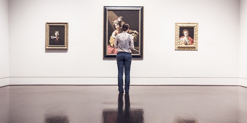 An image of a person standing in a museum, looking at three framed paintings.