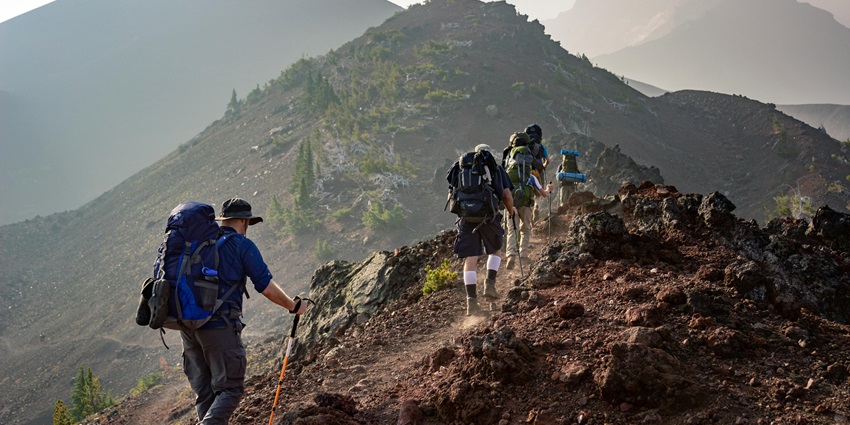 An image of Hikers with backpacks trek a steep, rocky mountain trail toward a hazy peak.