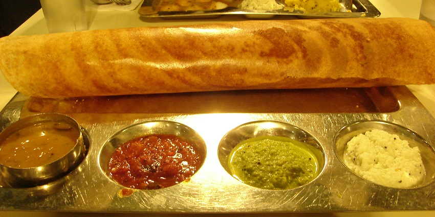 An image of Masala Dosa served with different chutneys and sambhar at a restaurant.