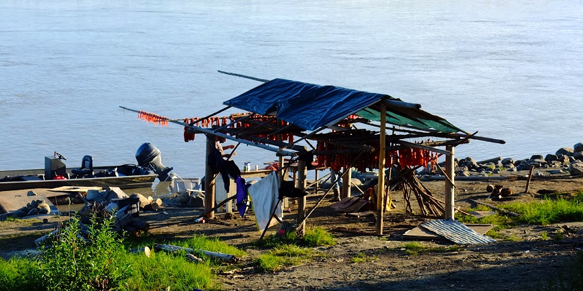 Fishing camp base near a lake and wood and much equipment laid near the grass and rocks.