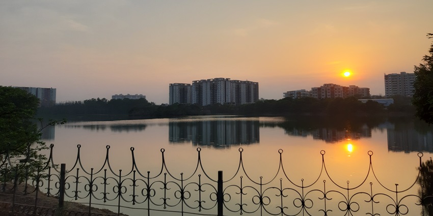 Sunset at Kaikondrahalli Lake with a still water body, railings and buildings in the back.