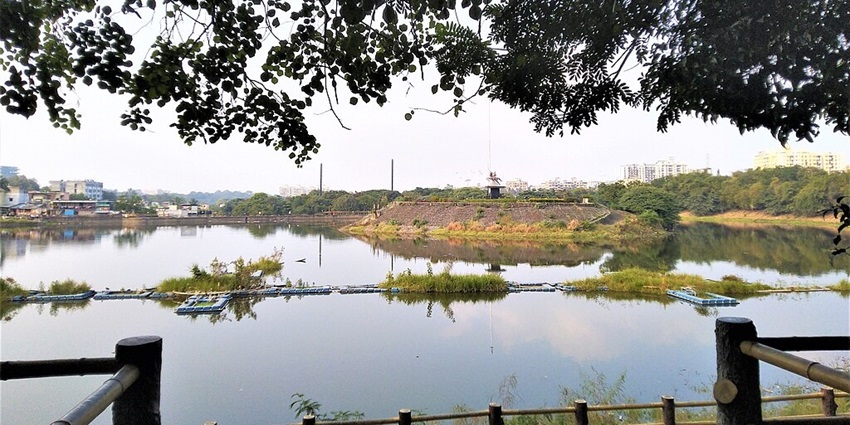 Lake surrounded by greenery and railings with buildings in the back of the waterbody
