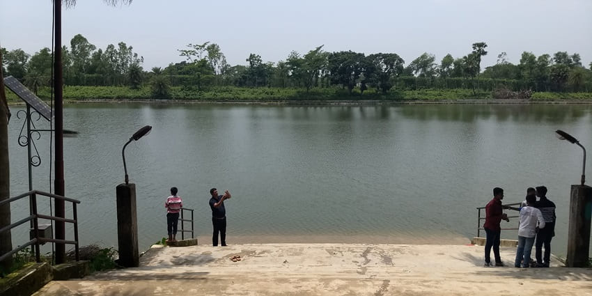 A man takes a photograph from his phone while standing near a lake on a sunny day.