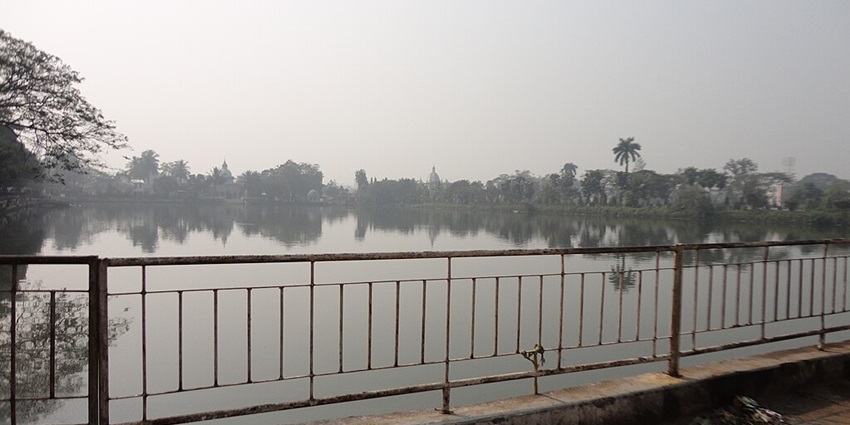 Lake in Agartala with railings, a distant temple surrounded by greenery and palm trees