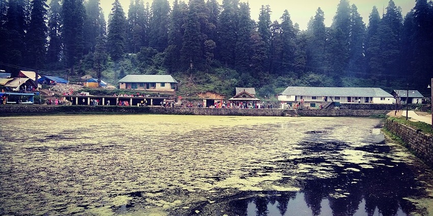 Kamrunag temple area with buildings and people beyond the lake covered in greenery.