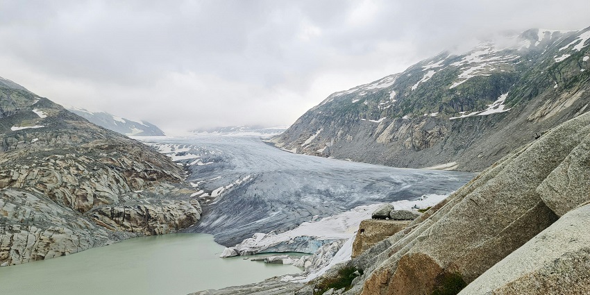 Lake beside a glacier surrounded by rocky mountains partially covered with snow .