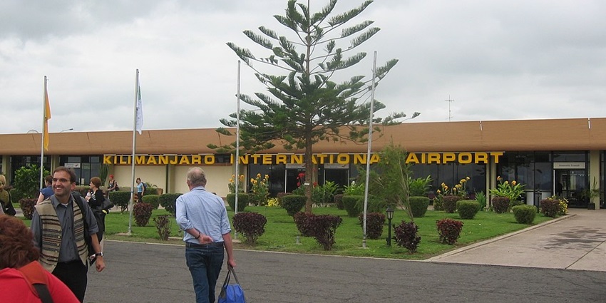 Entrance to the Kilimanjaro International Airport showing a brown building, bushes, plants, tree and people