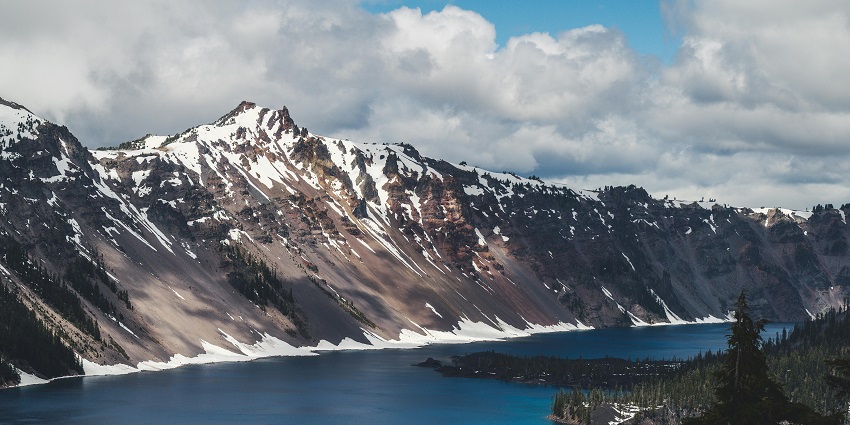 Dark blue lake with green trees on the right side and snowy grey mountains on the other side.