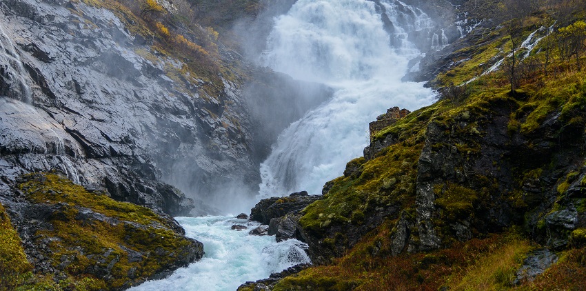 An Image of water flowing from the Kjosfossen Waterfall, surrounded by greenery and rocky cliffs.