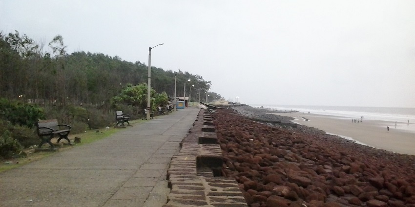 A paved promenade with benches runs alongside a rocky shoreline and a sandy beach