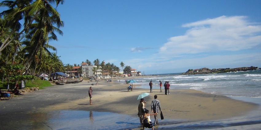 An image of Kovalam beach with people, palm trees, and waves under a clear blue sky