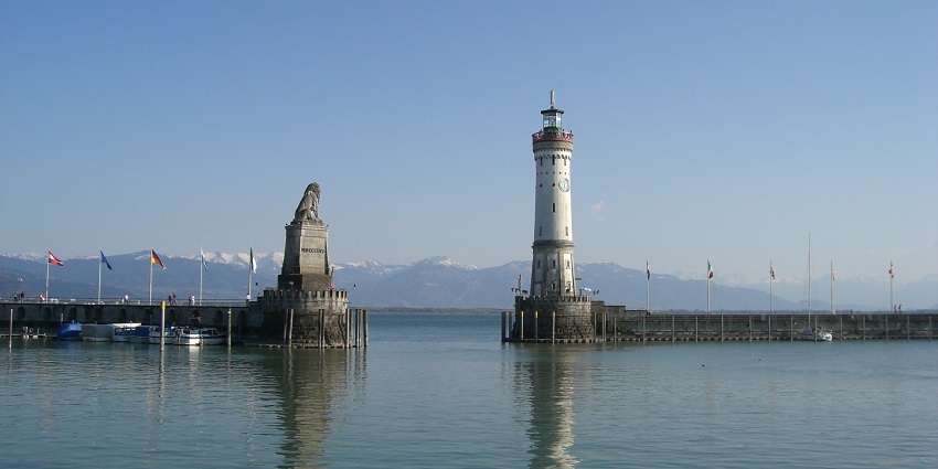 An image of Lake Constance with a lighthouse and a statue and distant mountains.